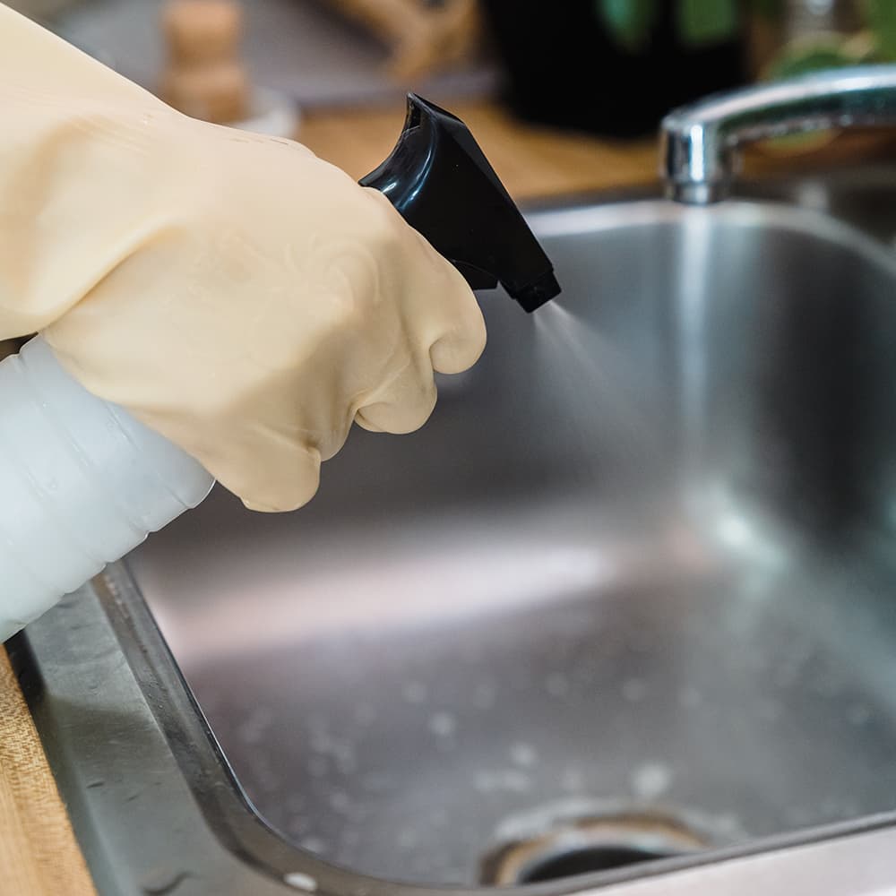 A hand wearing a yellow glove spraying disinfectant spray into a sink